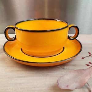 Op de afbeelding: Een helder oranje keramische soepkom met een zwarte rand en twee handvatten. De kom staat op een bijpassend schoteltje.