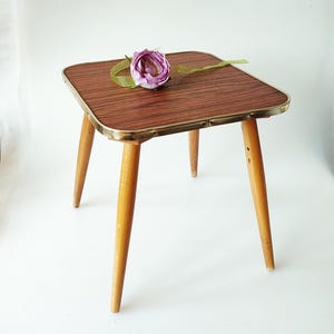 Puede incluir: Una pequeña mesa auxiliar cuadrada con tapa de imitación madera y ribete de latón. La mesa tiene cuatro patas de madera cónicas. Una flor morada y una cinta verde adornan la parte superior. La mesa mide unos 46 cm de alto.