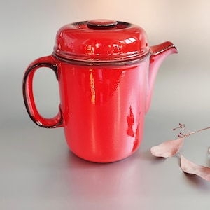 Puede incluir: Una cafetera de cerámica roja con tapa y asa negra. La cafetera tiene un acabado brillante.