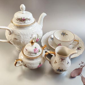 Può includere: Un set da tè in porcellana bianca con motivi floreali e dettagli dorati. Il set comprende una teiera, un creamer, uno zuccheriera, una tazza e un piattino.