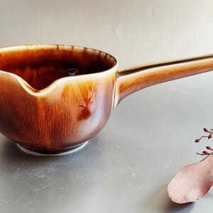 以下が含まれることがあります： 長い柄のついた茶色のセラミックソースポット。ポットには注ぎ口があり、光沢のある仕上がりです。ハンドルは明るい茶色です。このポットはおそらく、ソースの調理や提供に使用されます。