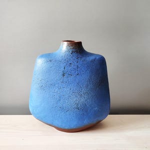 Op de afbeelding: Een blauwe keramische vaas met een getextureerd oppervlak. De vaas heeft een brede, afgeronde body en een smalle hals met een bruine rand. De basis is ook bruin. De vaas is een decoratief item.
