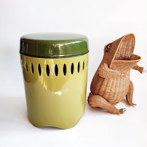 May include: A vintage olive green and yellow metal trash can with oval cutouts and a dark green lid. Beside it is a woven wicker frog-shaped basket with its mouth open.