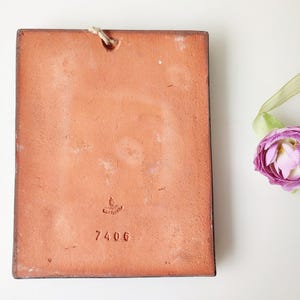 May include: A rectangular, terracotta-colored tile with a rough texture and a small hole at the top for hanging. The number "7406" is stamped on the tile. A purple flower with a green ribbon is in the background.