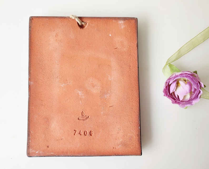May include: A rectangular, terracotta-colored tile with a rough texture and a small hole at the top for hanging. The number "7406" is stamped on the tile. A purple flower with a green ribbon is in the background.