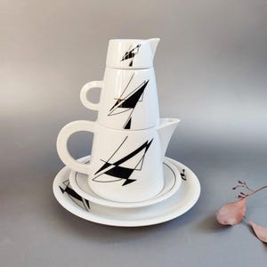 Puede incluir: Un juego de té de cerámica blanca compuesto por una tetera, una lechera y un azucarero apilados, rematados con una pequeña tapa. Cada pieza presenta un patrón de líneas geométricas negras. El conjunto descansa sobre un plato blanco a juego.