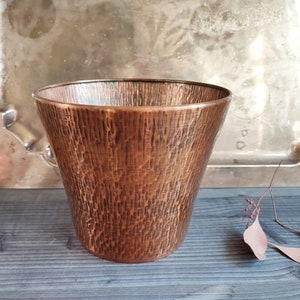 Könnte beinhalten: Ein gehämmerter Kupfertopf mit einem breiten, abgerundeten Boden und einem leicht ausgestellten Rand. Der Topf hat eine warme, rotbraune Farbe und eine strukturierte Oberfläche.