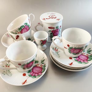 May include: A set of white teacups and saucers with pink and green floral designs. The set includes a small white egg cup and a white tea caddy with the word "Tee" written on the lid.