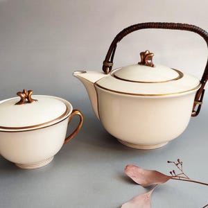 May include: A cream-coloured tea set with gold trim. The set includes a teapot with a woven handle and a lidded sugar bowl. The teapot and sugar bowl have gold accents on the lids and handles. The set is on a grey surface.