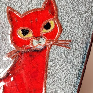 May include: A red cat illustration with yellow eyes and white whiskers. The cat is set against a textured, light gray background with a cracked pattern. The artwork has a handmade, folk art style.