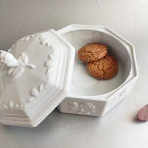 Könnte beinhalten: Weiße, achteckige Porzellanbox mit einem Deckel, der mit einem erhabenen Blumendesign verziert ist. Der Deckel ist geöffnet und die Box enthält zwei braune Kekse.