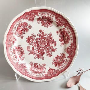 Puede incluir: Un plato de cena de cerámica blanca con un patrón floral rojo. El plato tiene un borde festoneado y un borde elevado.