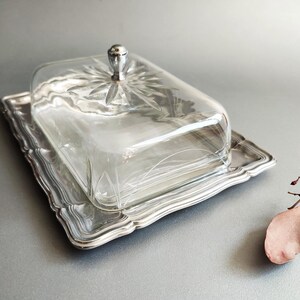 May include: A rectangular butter dish featuring a silver-toned metal tray with a scalloped edge and a clear glass lid. The glass cover is topped with a small, rounded silver-toned knob and displays a starburst pattern etched into the top surface.