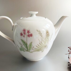 May include: White porcelain teapot with a floral design featuring pink flowers, green leaves, and brown stems. The teapot has a round body and a curved handle.