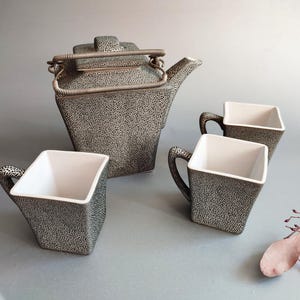 Op de afbeelding: Een keramische theeset met een theepot en drie vierkante kopjes. De set heeft een gespikkeld zwart-wit patroon aan de buitenkant, met witte interieurs. De theepot heeft een metalen handvat.