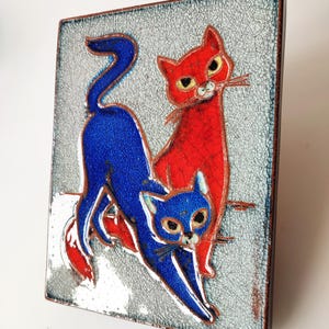May include: A rectangular ceramic tile featuring two stylized cats. One cat is red, the other is blue, set against a textured, light gray background. The tile has a dark brown border and is likely a decorative piece.
