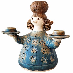 Può includere: Una statuetta in ceramica di una persona con cappello e capelli castani testurizzati, che indossa un abito blu decorato con un motivo a stelle ripetuto. La figura ha due braccia tese, ognuna delle quali sostiene un piccolo portacandele circolare.