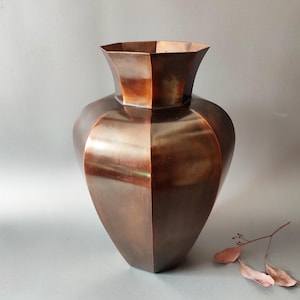 Könnte beinhalten: Eine große, kupferfarbene Vase mit geometrischem Design. Die Vase hat einen breiten, abgerundeten Körper und eine ausgestellte, facettierte Oberseite. Das Kupfer hat einen warmen, rötlich-braunen Farbton, und die Oberfläche reflektiert das Licht. Ein paar getrocknete Blätter liegen in der Nähe der Vase.