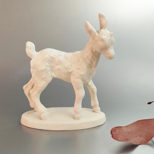 May include: A white ceramic figurine of a baby goat standing on a small oval base. The goat has a fluffy, textured coat and is posed with its head turned to the side.