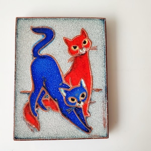 May include: A square, decorative tile featuring two stylized cats. One cat is bright red, the other is blue. The cats are set against a textured, light gray background. The tile has a dark brown border.