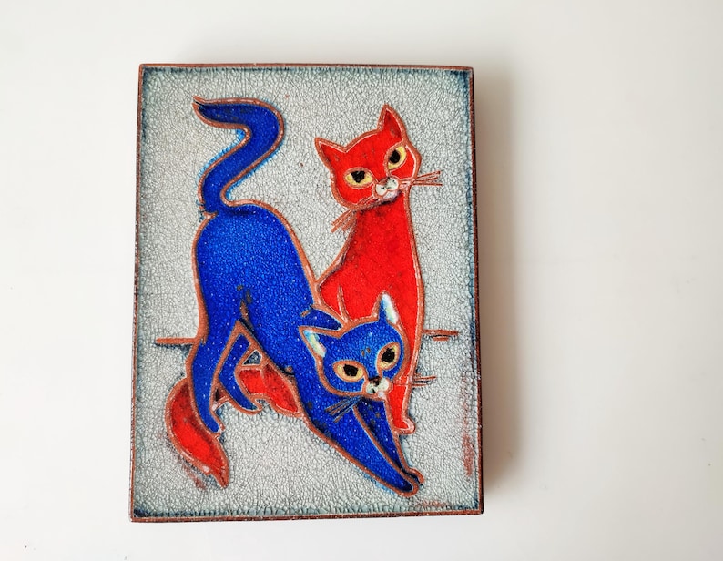 May include: A square, decorative tile featuring two stylized cats. One cat is bright red, the other is blue. The cats are set against a textured, light gray background. The tile has a dark brown border.