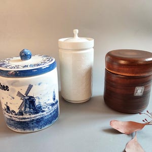 Puede incluir: Tres tarros de cerámica decorativos. Uno es blanco con tapa y asa pequeña. Otro es un tarro azul y blanco con una escena de molino de viento y la palabra "Thee". El tercero es un tarro de madera marrón.
