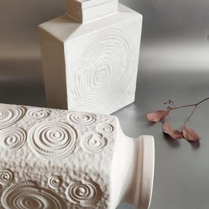 Puede incluir: Dos jarrones de cerámica blanca con un patrón de espiral. El jarrón de la izquierda es cilíndrico y el jarrón de la derecha es cuadrado.
