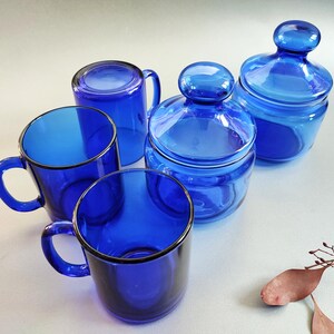 May include: A collection of cobalt blue glass drinkware and storage jars. The set includes two handled mugs, a mug without a handle, and two jars with lids. The glass has a glossy finish and a dark blue color.