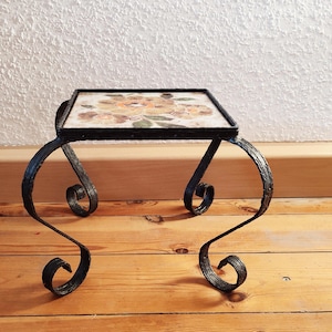 May include: A small black metal table with a floral tile top. The table has a scrolled design on the legs.
