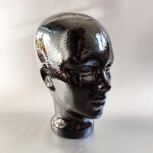 May include: A glossy, dark brown ceramic head bust with a textured surface. The bust features detailed facial features, including a nose, ears, and eyes. The head is mounted on a circular base. The bust reflects light, creating highlights and shadows.