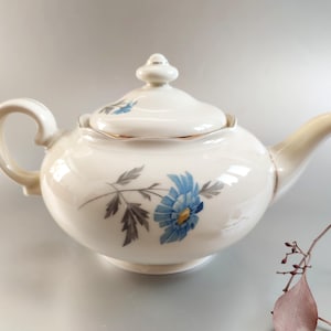 Puede incluir: Una tetera de porcelana blanca con un diseño floral azul. La tetera tiene una tapa y un asa. El diseño floral presenta una flor azul con un centro amarillo y hojas grises.