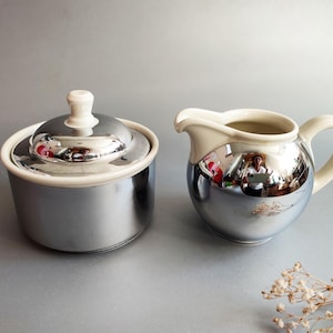 May include: A ceramic sugar bowl with a lid and a matching creamer pitcher. Both pieces feature a reflective, mirror-like silver metallic finish on the lower sections, contrasted with a smooth, off-white cream-colored ceramic top and handle.