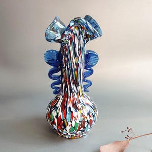 Puede incluir: Un jarrón de cristal artístico colorido con base bulbosa y parte superior acampanada. El jarrón presenta un patrón moteado de rojo, blanco, azul y verde. Tiene detalles azules y un borde con volantes, agregando un toque decorativo.