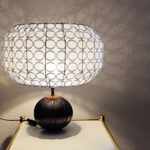 Puede incluir: Una pantalla de lámpara blanca con un patrón geométrico azul y blanco se asienta sobre una base marrón oscuro. La lámpara está sobre una mesa blanca con un borde dorado.