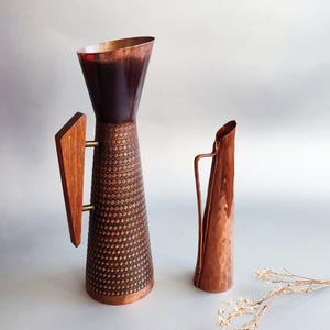 Könnte beinhalten: Ein hoher Kupferkrug mit texturiertem Rautenmuster und dreieckigem Holzgriff, gepaart mit einem kleineren, schlanken Kupferkrug mit gehämmertem Finish und dünnem Metallgriff, daneben ein kleiner Zweig mit getrockneten weißen Blumen.