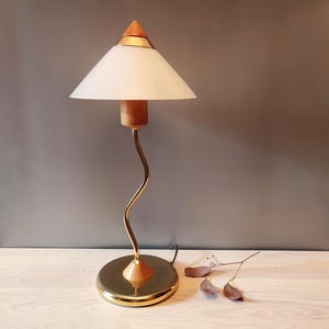 Puede incluir: Una lámpara de mesa vintage con base de latón y un tallo curvo en tono dorado. La lámpara presenta detalles en madera y una pantalla cónica blanca. La parte superior de la lámpara tiene forma de cono de madera. La lámpara está sobre una superficie de madera clara.