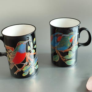 May include: Two black ceramic mugs with colorful bird illustrations. One mug features a red, blue, and green bird with a black background. The other mug features two blue and green birds with a black background.