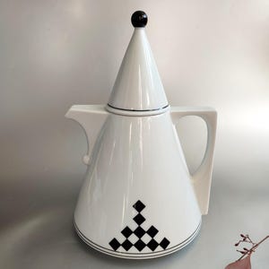 May include: White ceramic teapot with a conical lid and a black finial. The teapot features a geometric black and white diamond pattern near the base and a black line detail. The handle is square and the spout is curved.