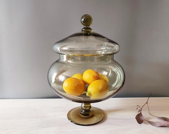 May include: A large, clear glass jar with a lid, filled with several bright yellow lemons. The jar has a wide, rounded body and a pedestal base. The glass has a slight smoky tint.