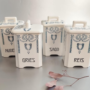 Könnte beinhalten: Vier weiße Keramik-Gewürzgläser mit Deckeln und Griffen. Jedes Glas hat eine rechteckige Form und ist mit blauen floralen Designs und Text versehen. Die Gläser sind mit "NUDEL", "GRIES", "SAGO" und "REIS" beschriftet.