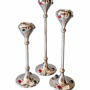 Puede incluir: Tres candelabros de metal en tono plateado de diferentes alturas, cada uno con un tallo delgado, una base acampanada y una parte superior redondeada. Las superficies están adornadas con pequeñas piedras cabujón circulares incrustadas en colores rojo, verde y azul.