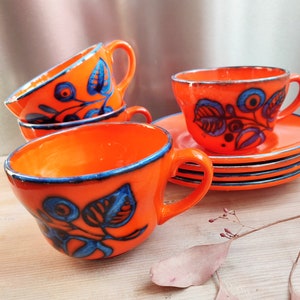 May include: Set of four orange ceramic teacups with blue and purple floral designs. The cups have a blue rim and are stacked on top of each other. The cups are on a wooden surface with a dried leaf.