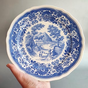 Puede incluir: Un plato redondo de cerámica con borde festoneado que presenta un patrón de transferencia azul y blanco. El centro representa un paisaje con un río, árboles y figuras, mientras que el borde está decorado con motivos florales y volutas.