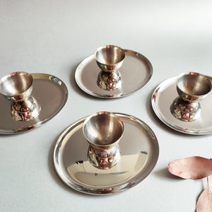 May include: A set of four polished stainless steel egg cups, each featuring a small, rounded bowl centered on a flat, circular plate base. The metallic surface is reflective, showing a smooth, clean finish across all four individual pieces.