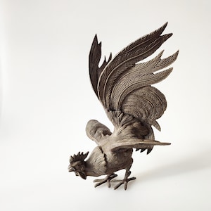 Puede incluir: Una figura de gallo detallada de color plateado con las alas extendidas. El gallo está de pie sobre una superficie blanca.