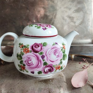 May include: A white teapot with a floral design featuring pink and purple roses, orange and yellow flowers, and green leaves. The teapot has a lid with a red trim.