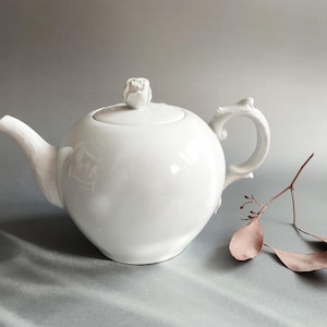 May include: A white ceramic teapot with a rounded body, a curved spout, and a handle. The lid features a small sculpted rose as a knob. Beside the teapot lies a thin, brown, dried twig with several small, elongated, dried leaves attached to it.