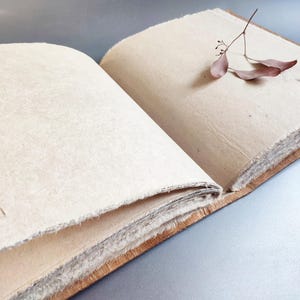 May include: An open, handmade journal with thick, off-white pages and a brown leather binding. A small sprig of dried leaves rests on one page. The paper has a textured, deckled edge.