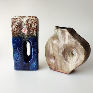 Op de afbeelding: Twee keramische vazen, een met een blauwe en bruine glazuur en een rechthoekige vorm met een gat in het midden, de andere met een lichtbruine glazuur en een onregelmatige vorm met een gat in het midden.