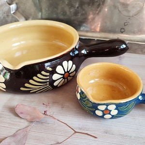 May include: Two small ceramic bowls with handles. The larger bowl is brown with yellow and white daisy designs. The smaller bowl is blue with green and white daisy designs.
